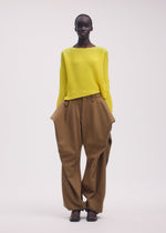 A person wears the ISSEY MIYAKE SHIFTED MISTY PLEATS: a striking yellow long-sleeved top with unique design, paired with loose brown pants featuring wide, structured hips and dark shoes, standing against a plain background.