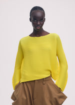 A woman with braided hair models the ISSEY MIYAKE SHIFTED MISTY PLEATS bright yellow long-sleeved top and tan high-waisted trousers, standing against a plain light background with her hands in her pockets.