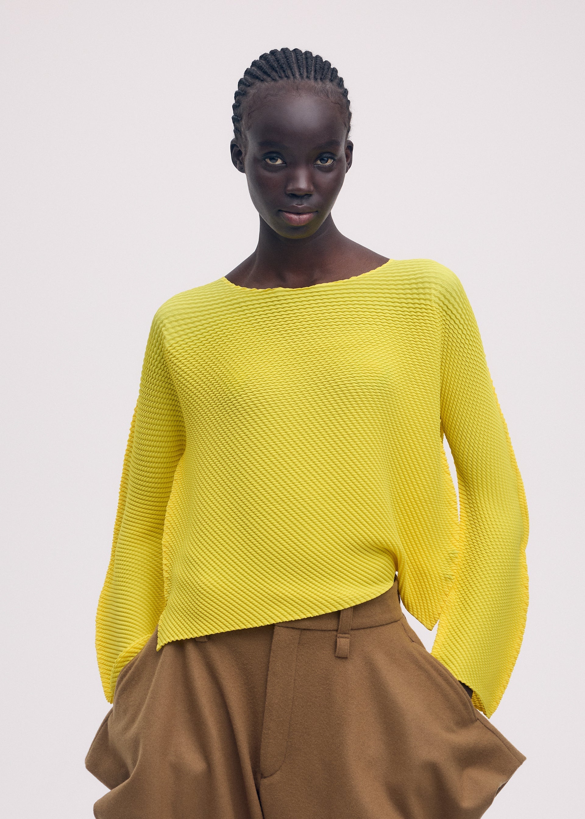 Zoomed view A woman with braided hair models the ISSEY MIYAKE SHIFTED MISTY PLEATS bright yellow long-sleeved top and tan high-waisted trousers, standing against a plain light background with her hands in her pockets.