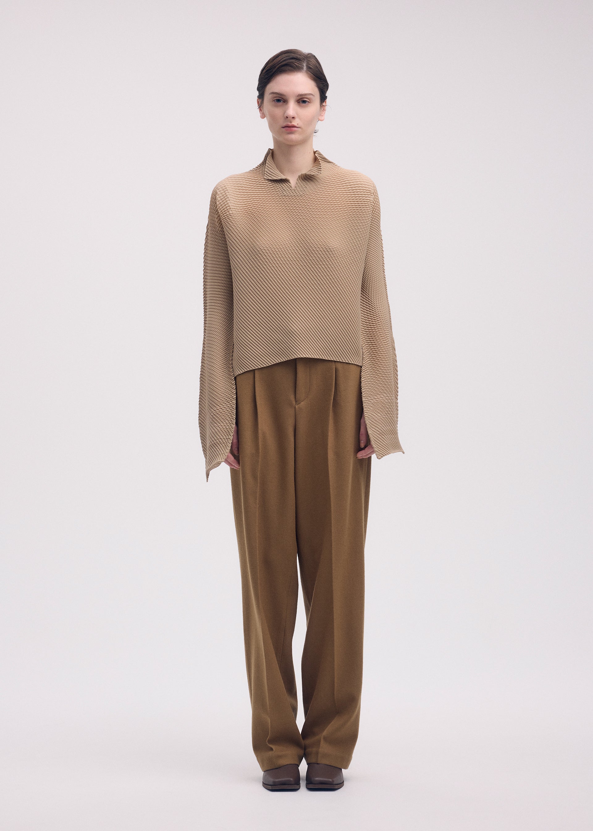 Zoomed view A woman faces forward in ISSEY MIYAKE's SHIFTED MISTY PLEATS: a loose, textured beige pleated top with long sleeves, paired with wide-legged, high-waisted brown trousers. Her dark hair is pulled back against a plain light background.