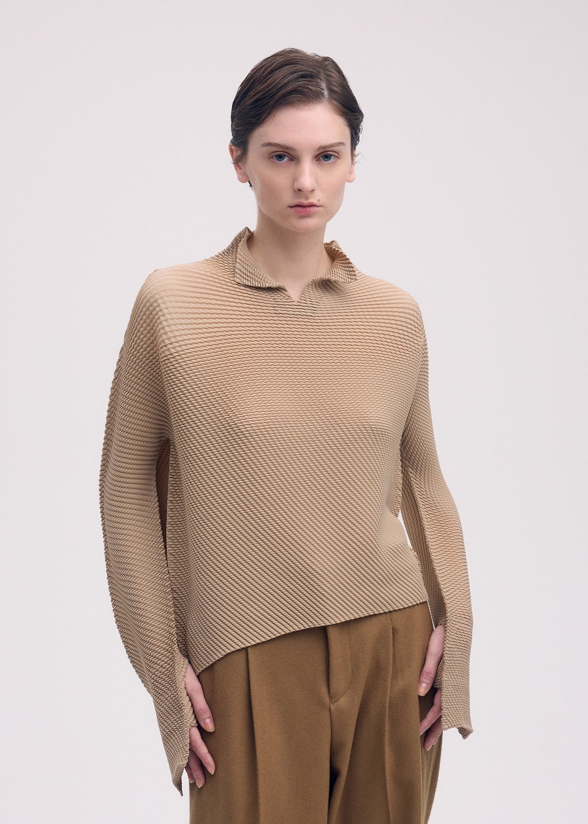A person with short brown hair models the ISSEY MIYAKE SHIFTED MISTY PLEATS textured beige long-sleeved top and brown trousers, standing against a plain light background with a neutral expression. view 6 of 7