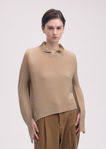 A person with short brown hair models the ISSEY MIYAKE SHIFTED MISTY PLEATS textured beige long-sleeved top and brown trousers, standing against a plain light background with a neutral expression.