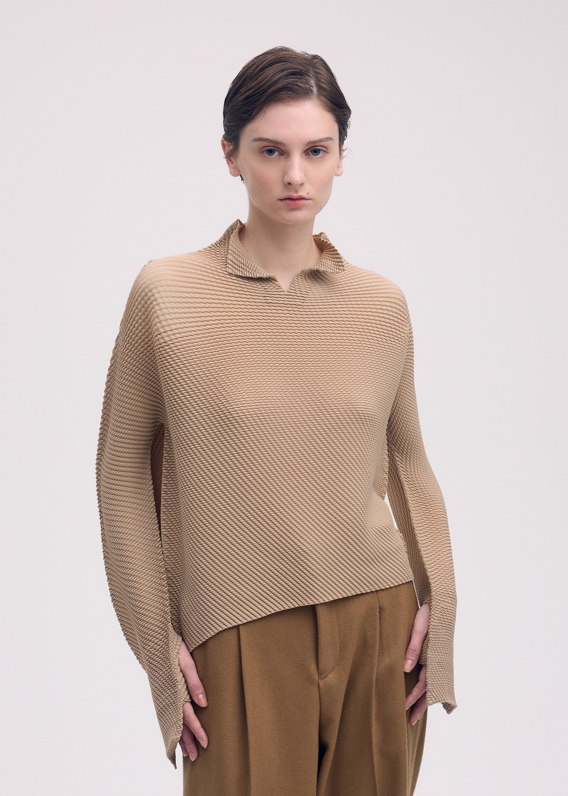 Zoomed view A person with short brown hair models the ISSEY MIYAKE SHIFTED MISTY PLEATS textured beige long-sleeved top and brown trousers, standing against a plain light background with a neutral expression.