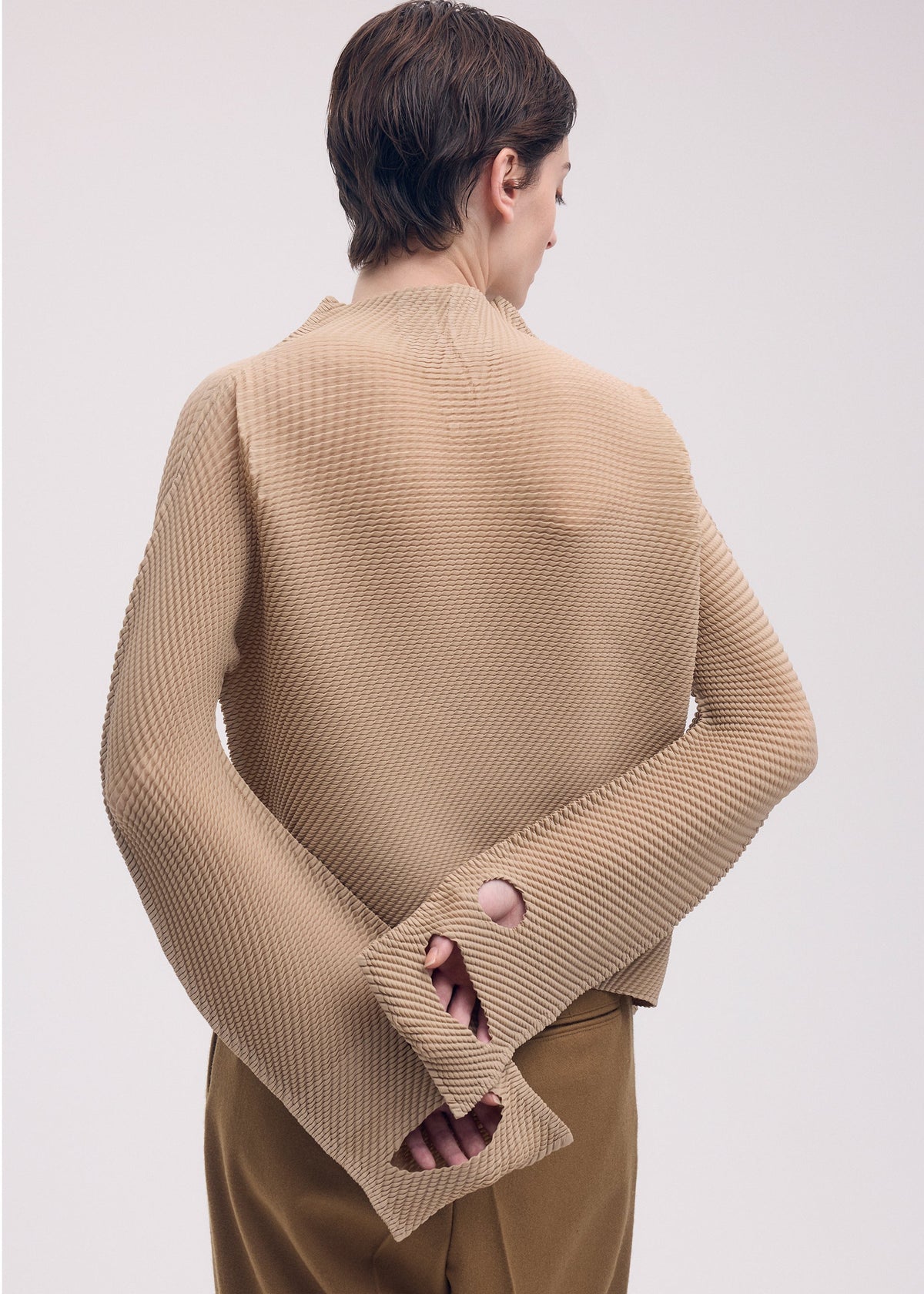A person with short brown hair stands facing away, wearing ISSEY MIYAKE's SHIFTED MISTY PLEATS textured long-sleeved top with oversized sleeves and wrist holes, paired with brown pants, set against a light background. view 7 of 7