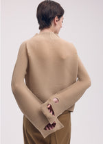 A person with short brown hair stands facing away, wearing ISSEY MIYAKE's SHIFTED MISTY PLEATS textured long-sleeved top with oversized sleeves and wrist holes, paired with brown pants, set against a light background.