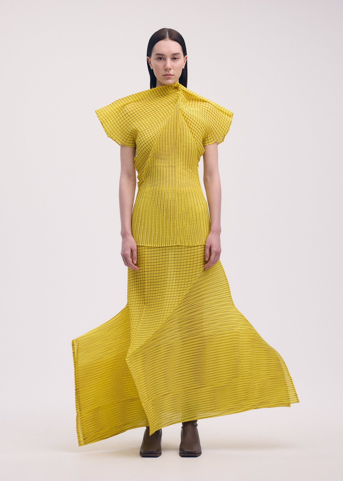 A woman with straight dark hair and a neutral expression wears ISSEY MIYAKE’s GENERIC STRIPES (MACHINE PLEATS) yellow striped pleated top with geometric lines, paired with brown ankle boots, against a plain background. view 5 of 7