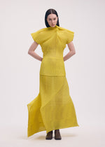 A woman stands against a plain background wearing the ISSEY MIYAKE GENERIC STRIPES (MACHINE PLEATS) set—a bright yellow, striped pleated top with angular shoulders and a matching long skirt—paired with brown boots. Her dark hair is center-parted and straight.