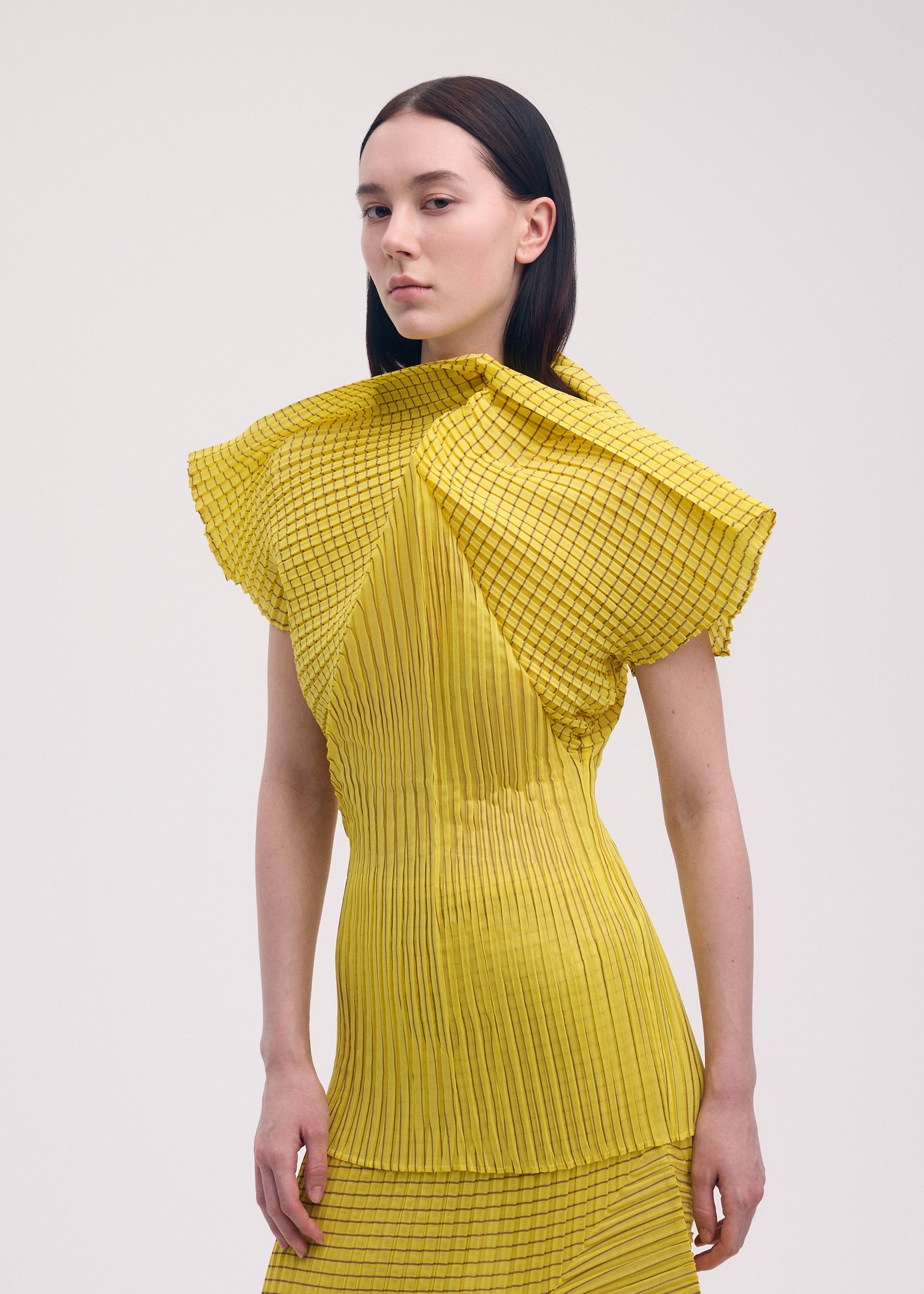 Zoomed view A person with straight dark hair wears the ISSEY MIYAKE GENERIC STRIPES (MACHINE PLEATS) dress, featuring pleated yellow fabric with black grid-like stripes, standing against a plain light background.