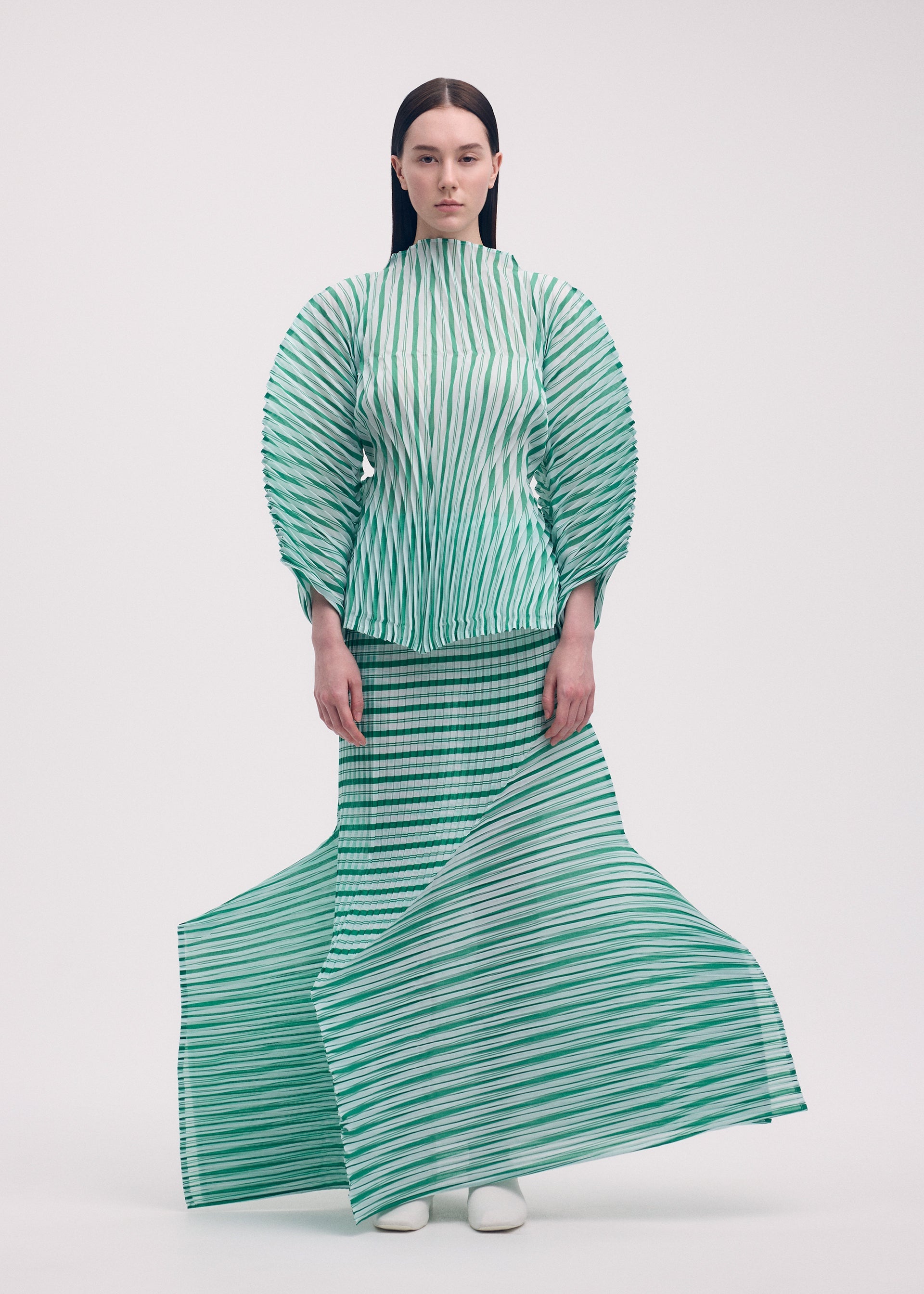 Zoomed view A woman faces forward in ISSEY MIYAKE's GENERIC STRIPES (HAND PLEATS) set: a striped pleated top with voluminous sleeves and matching flowing skirt. She has straight dark hair, white shoes, and stands against a plain white background.