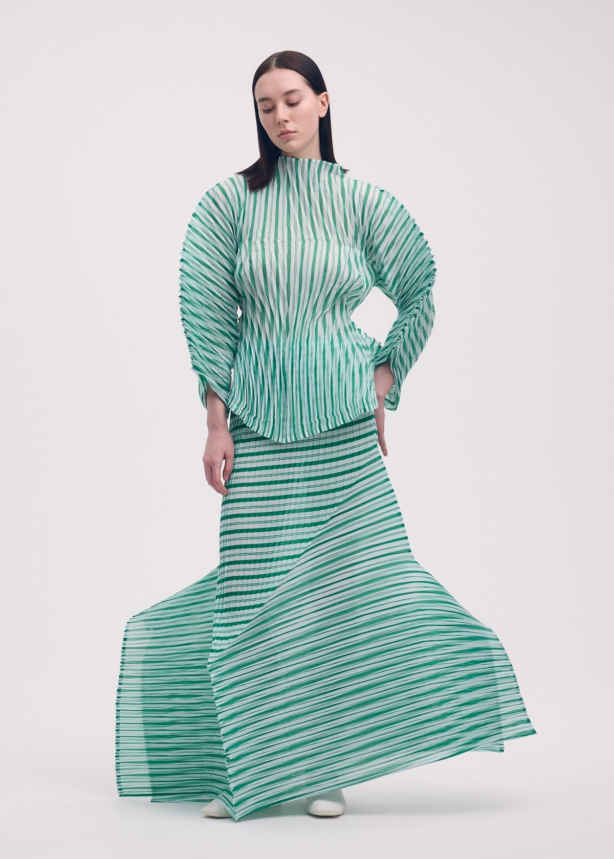 A woman with long, straight dark hair stands against a plain background, wearing ISSEY MIYAKE’s GENERIC STRIPES (HAND PLEATS) set—green and white striped pleated top with matching skirt that flares dynamically at the hem. view 4 of 5