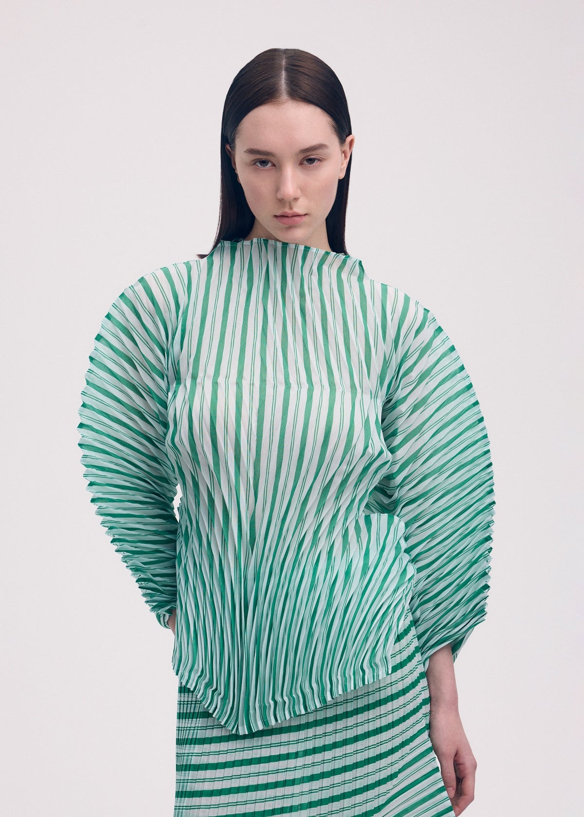 A woman with straight brown hair wears the ISSEY MIYAKE GENERIC STRIPES (HAND PLEATS) green and white striped pleated top with voluminous sleeves and matching skirt, standing against a plain light background. view 5 of 5