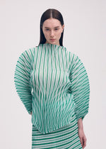 A woman with straight brown hair wears the ISSEY MIYAKE GENERIC STRIPES (HAND PLEATS) green and white striped pleated top with voluminous sleeves and matching skirt, standing against a plain light background.