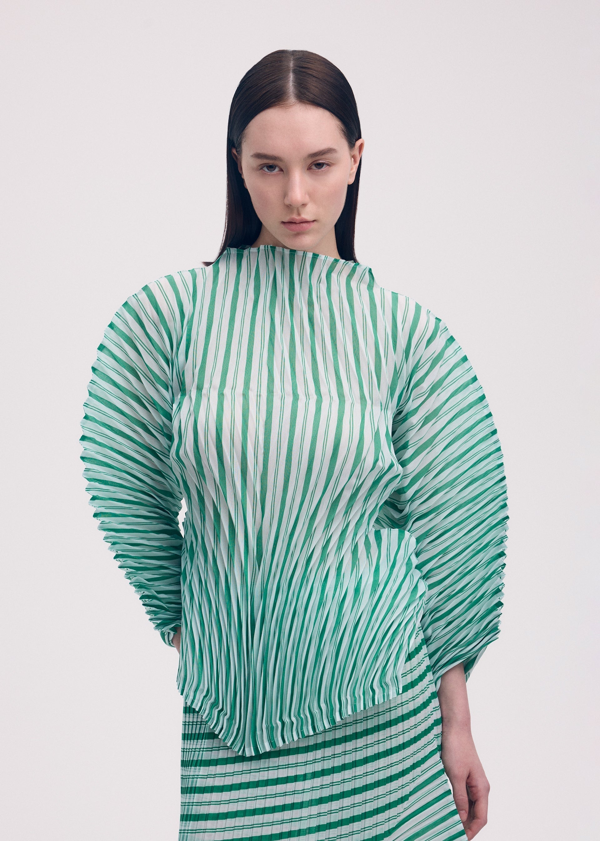 Zoomed view A woman with straight brown hair wears the ISSEY MIYAKE GENERIC STRIPES (HAND PLEATS) green and white striped pleated top with voluminous sleeves and matching skirt, standing against a plain light background.