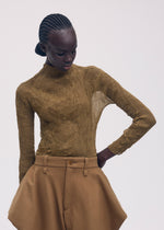 A person with braided hair models the ISSEY MIYAKE CHIFFON TWIST 3, a sheer high-neck olive-green long-sleeve top paired with high-waisted tan wide-leg pants, posing against a plain light backdrop with one hand on their hip.