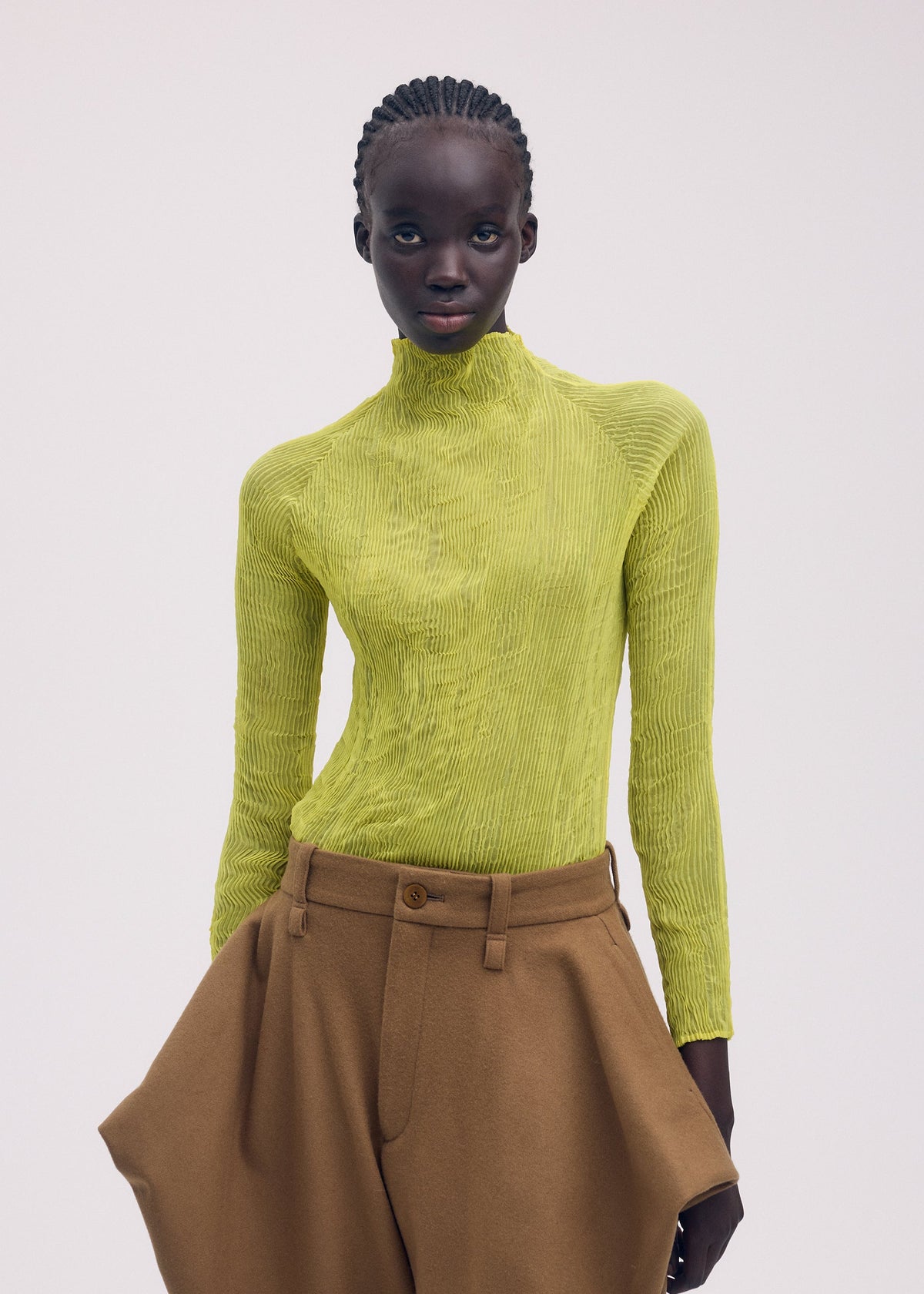 A person with neat cornrows poses against a plain light background, wearing ISSEY MIYAKE's CHIFFON TWIST 3—a bright yellow pleated top made from recycled polyester—and high-waisted, loose-fitting brown pants. view 7 of 7