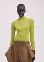 A person with neat cornrows poses against a plain light background, wearing ISSEY MIYAKE's CHIFFON TWIST 3—a bright yellow pleated top made from recycled polyester—and high-waisted, loose-fitting brown pants.