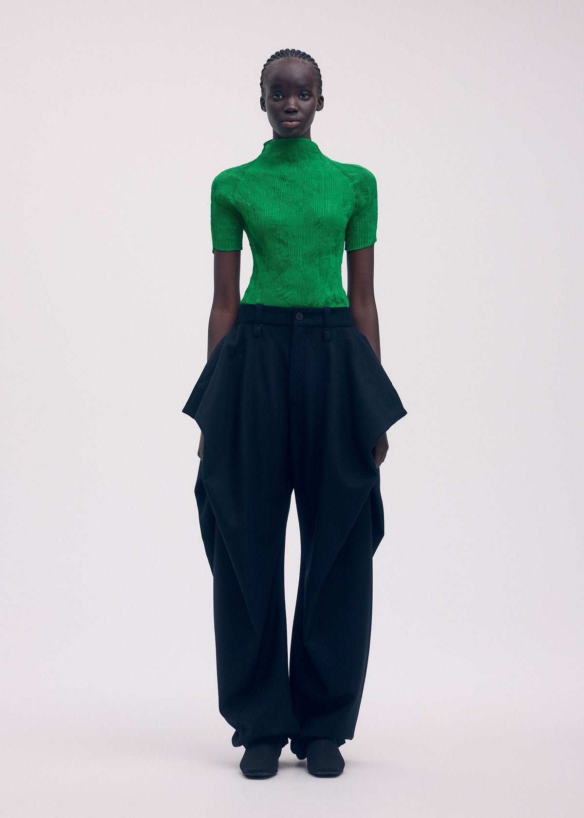 A person stands facing forward, wearing the ISSEY MIYAKE CHIFFON TWIST 3: a bright green recycled polyester turtleneck top and high-waisted, wide-leg black pants with draped details, set against a plain light background. view 5 of 7
