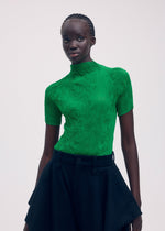 A person with dark skin and braided hair wears ISSEY MIYAKE’s CHIFFON TWIST 3, a bright green pleated top made from recycled polyester, paired with a high-waisted black skirt against a plain light background.