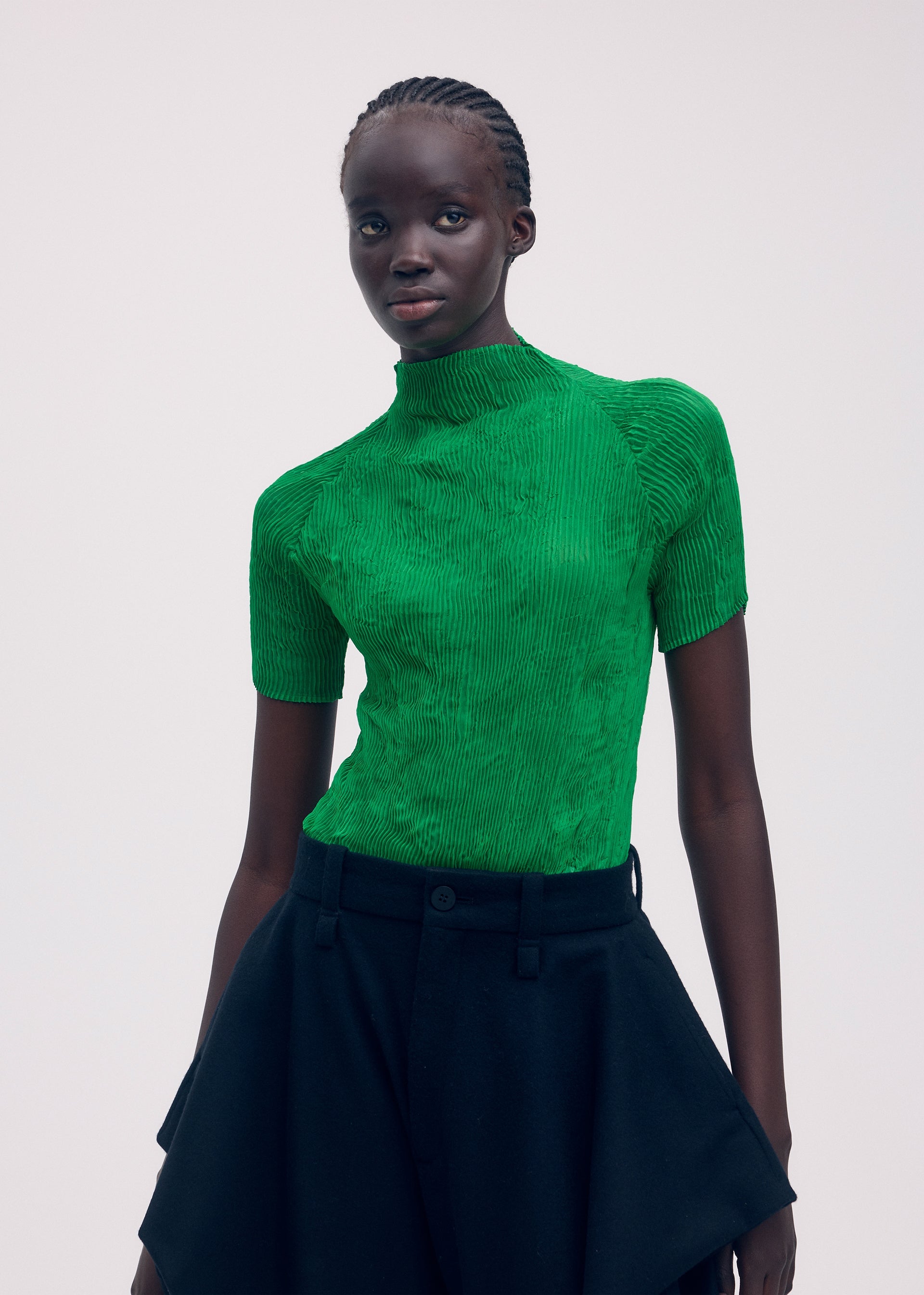 Zoomed view A person with dark skin and braided hair wears ISSEY MIYAKE’s CHIFFON TWIST 3, a bright green pleated top made from recycled polyester, paired with a high-waisted black skirt against a plain light background.