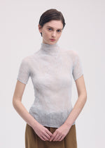 A person with short brown hair models the ISSEY MIYAKE CHIFFON TWIST 3—a sheer, light-toned, pleated recycled polyester top—paired with brown pants and stands against a plain white background, hands loosely clasped.