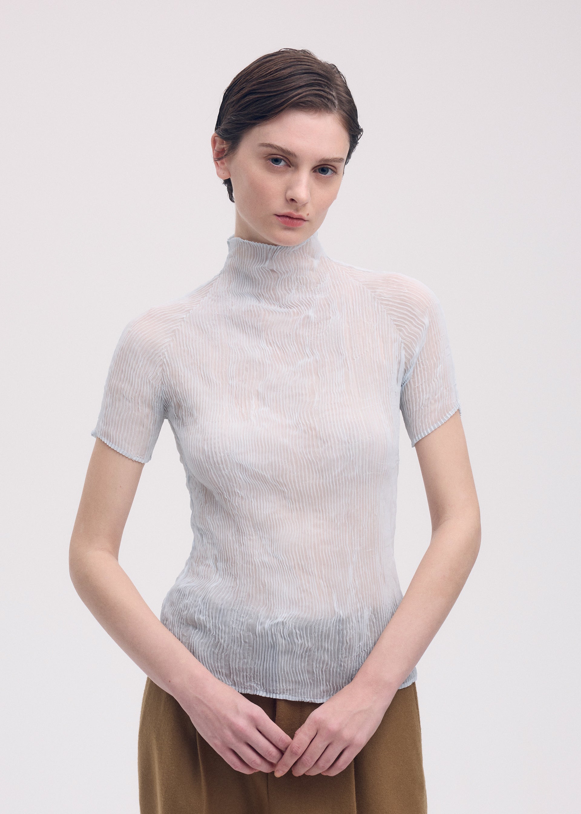 Zoomed view A person with short brown hair models the ISSEY MIYAKE CHIFFON TWIST 3—a sheer, light-toned, pleated recycled polyester top—paired with brown pants and stands against a plain white background, hands loosely clasped.