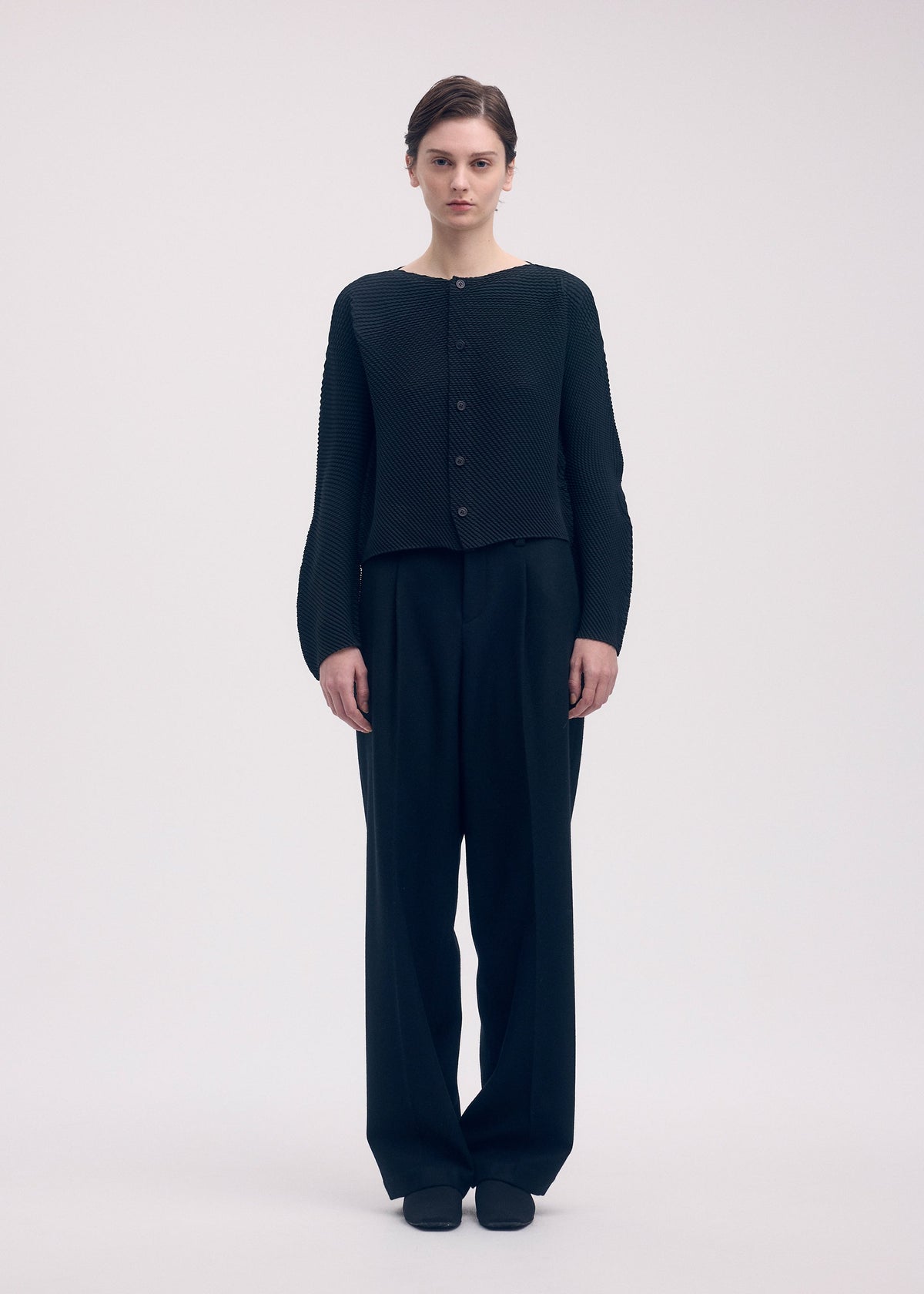 A person stands against a plain background, wearing ISSEY MIYAKE's SHIFTED MISTY PLEATS pleated black cardigan and loose black trousers, arms relaxed at their sides. view 5 of 6