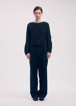 A person stands against a plain background, wearing ISSEY MIYAKE's SHIFTED MISTY PLEATS pleated black cardigan and loose black trousers, arms relaxed at their sides.