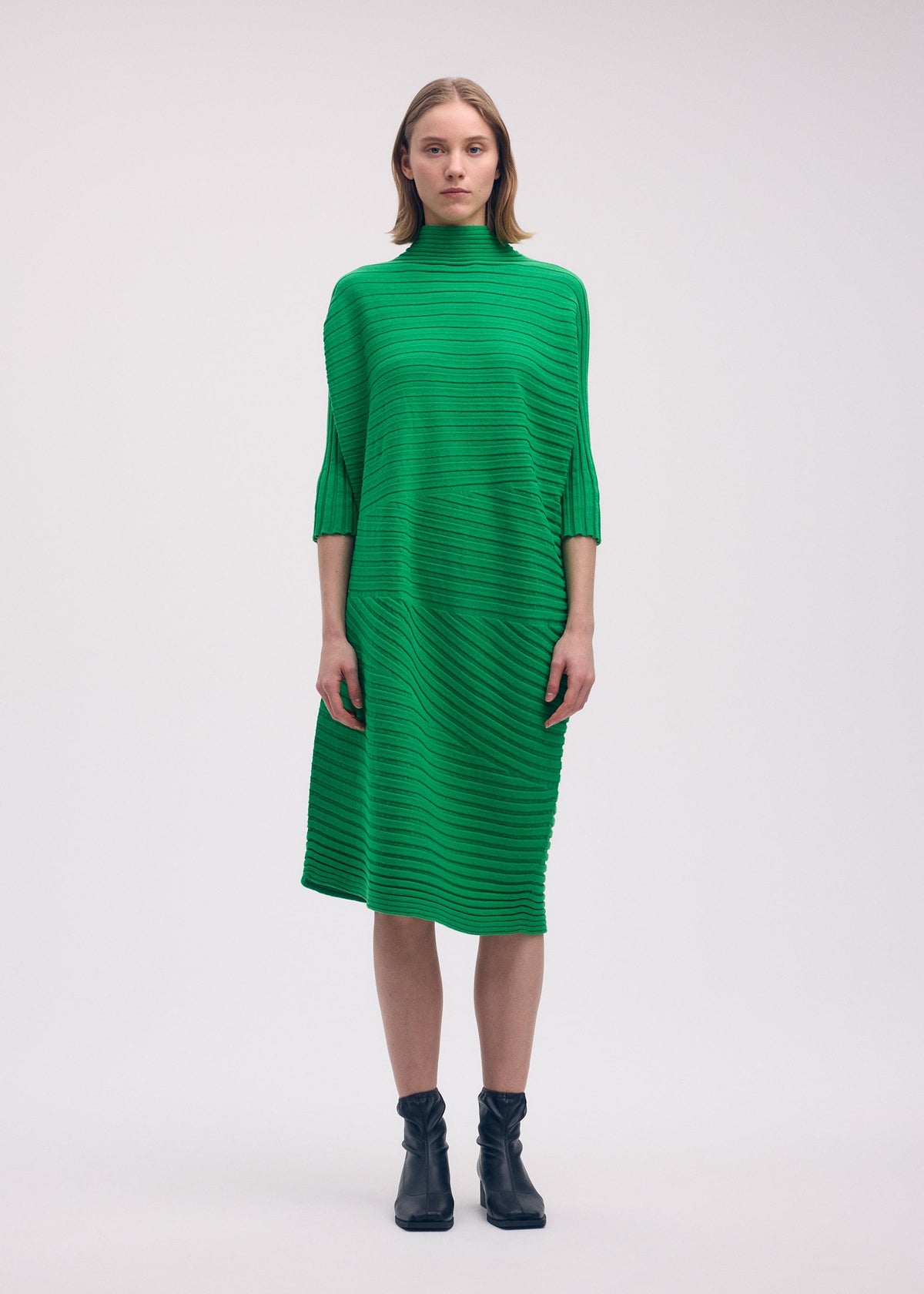 A woman faces forward in a knee-length, bright green COTTON CASHMERE KNIT dress by ISSEY MIYAKE, featuring a straight silhouette and half sleeves. She pairs it with black ankle boots; her straight hair is shoulder-length. White background. view 4 of 5