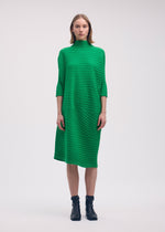 A woman faces forward in a knee-length, bright green COTTON CASHMERE KNIT dress by ISSEY MIYAKE, featuring a straight silhouette and half sleeves. She pairs it with black ankle boots; her straight hair is shoulder-length. White background.