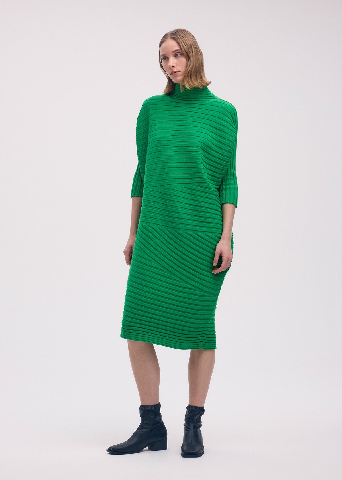 A woman with short hair stands before a plain light background in the ISSEY MIYAKE COTTON CASHMERE KNIT, a bright green knee-length dress with a high neckline and three-quarter sleeves, styled with black ankle boots. view 5 of 5