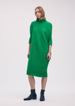 A woman with short hair stands before a plain light background in the ISSEY MIYAKE COTTON CASHMERE KNIT, a bright green knee-length dress with a high neckline and three-quarter sleeves, styled with black ankle boots.