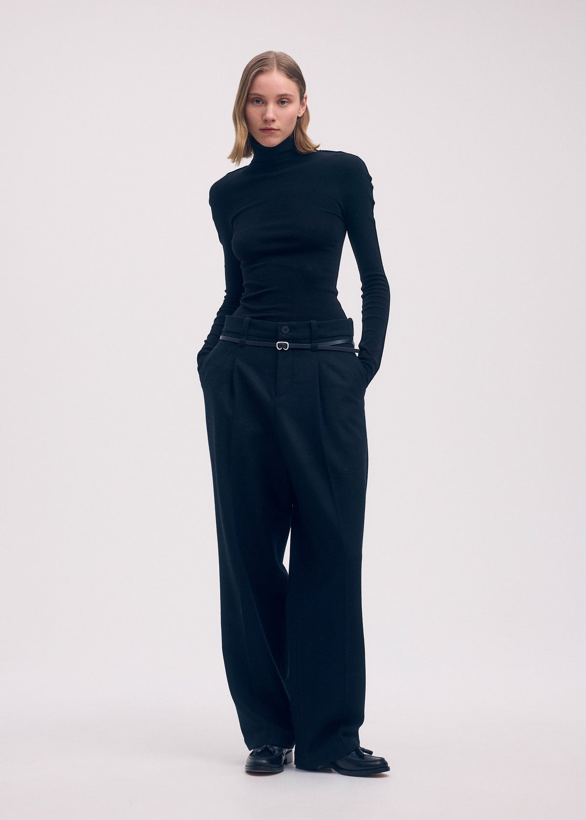 A person stands against a plain background holding the ISSEY MIYAKE COTTON BAGUETTE BK-58, wearing black attire with hands in their pockets and a neutral expression. view 4 of 4