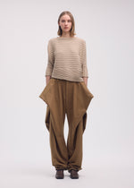 A woman with straight, shoulder-length hair and a neutral expression wears the ISSEY MIYAKE COTTON CASHMERE KNIT sweater in beige and loose brown pants with angular pockets, standing against a plain background.