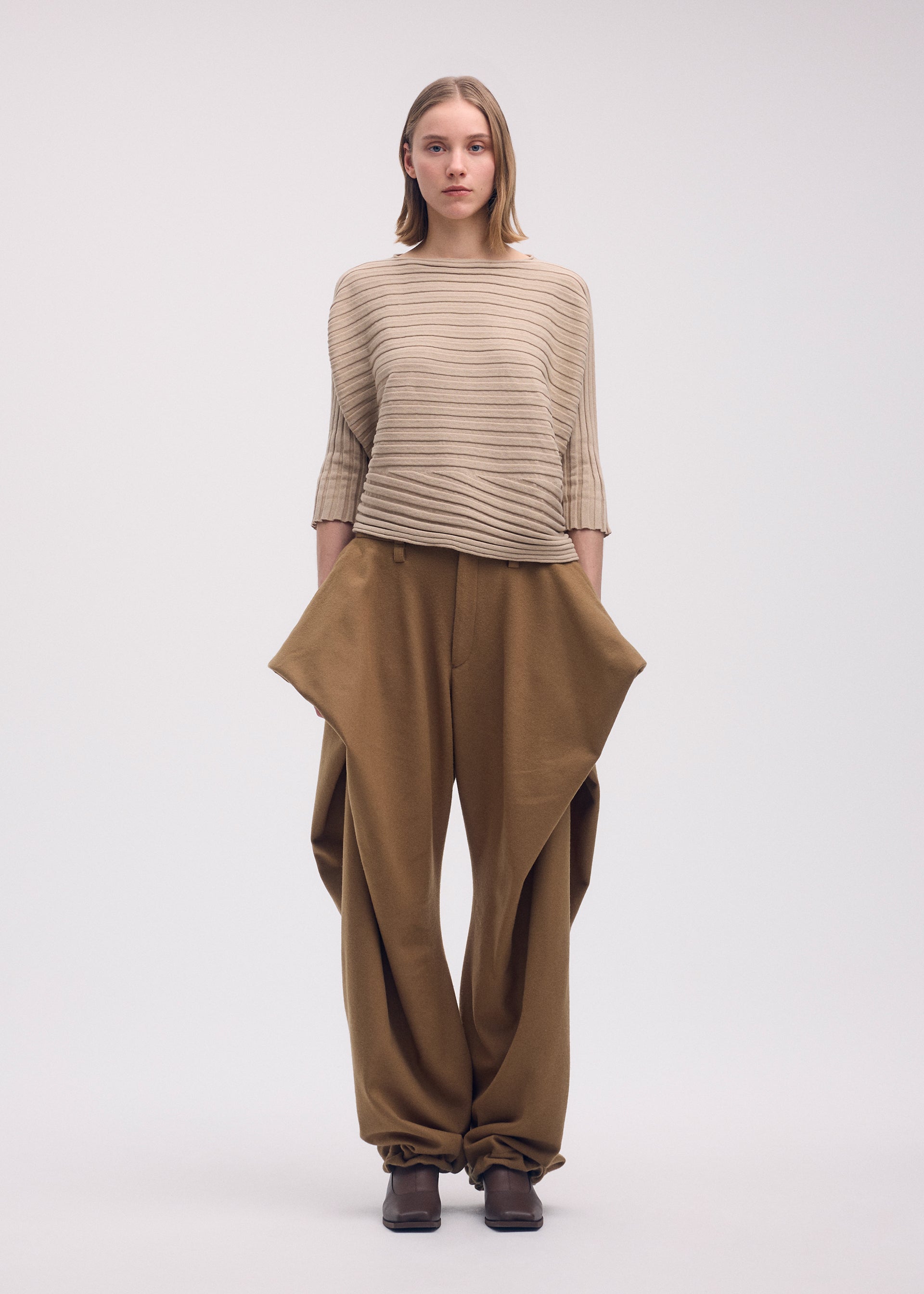 Zoomed view A woman with straight, shoulder-length hair and a neutral expression wears the ISSEY MIYAKE COTTON CASHMERE KNIT sweater in beige and loose brown pants with angular pockets, standing against a plain background.