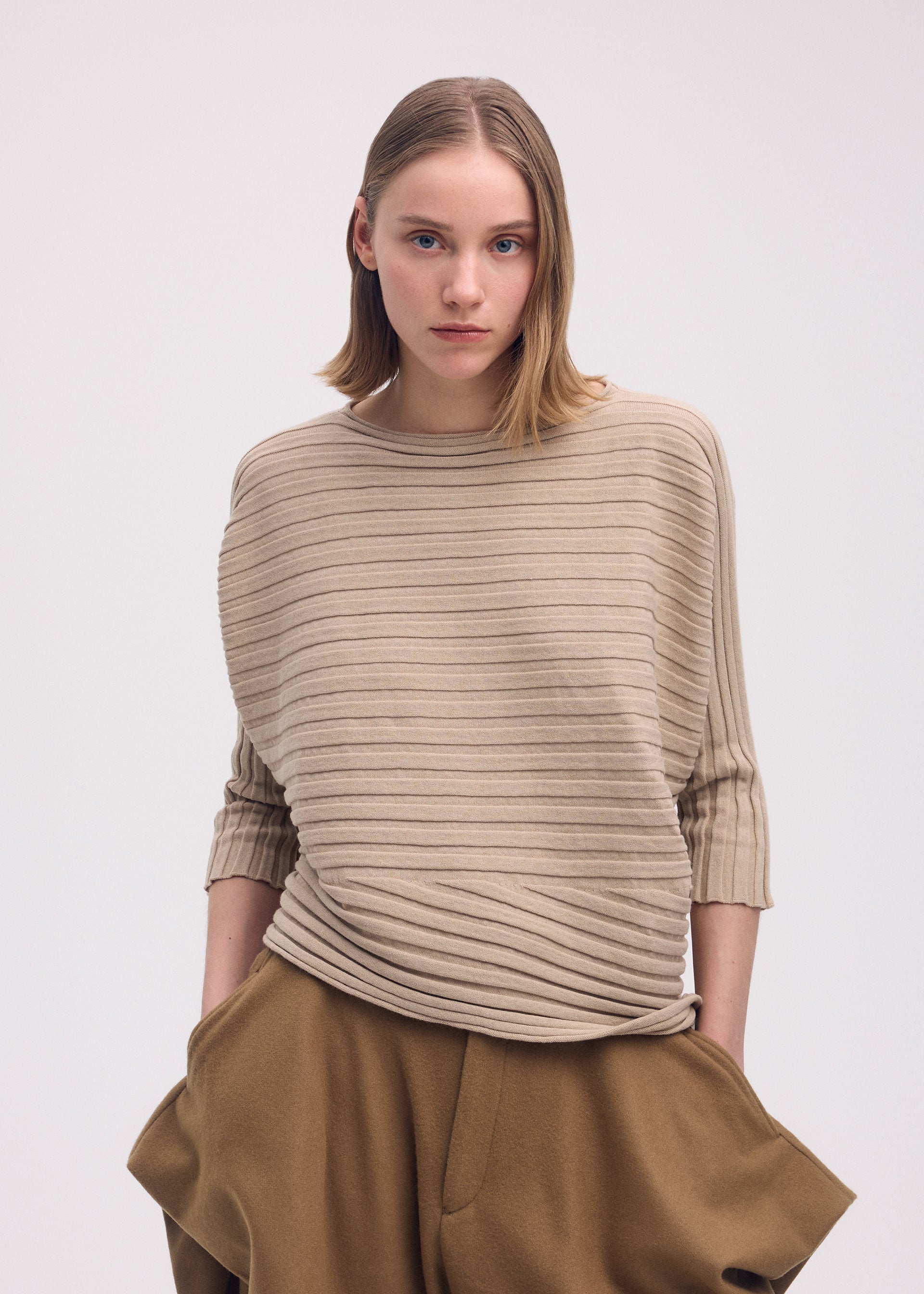 Zoomed view A young woman with straight, shoulder-length hair wears the ISSEY MIYAKE COTTON CASHMERE KNIT sweater in textured beige and loose brown pants, standing with her hands in her pockets against a plain light background.