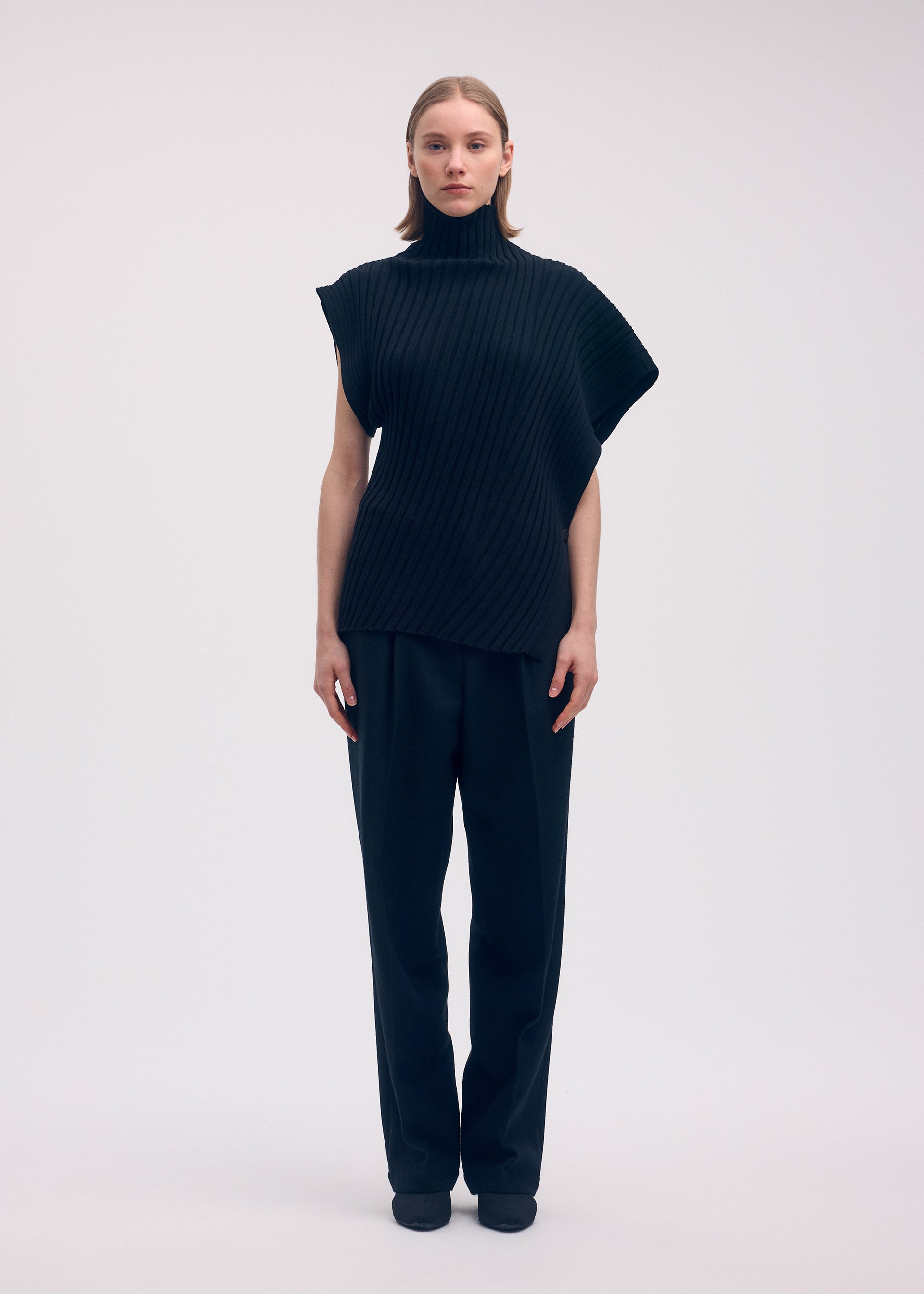 Zoomed view Against a plain background, a person faces forward in ISSEY MIYAKE's COTTON CASHMERE KNIT—a dark, ribbed high-neck top with asymmetrical shoulders—paired with loose dark trousers for a straight silhouette and a neutral expression.
