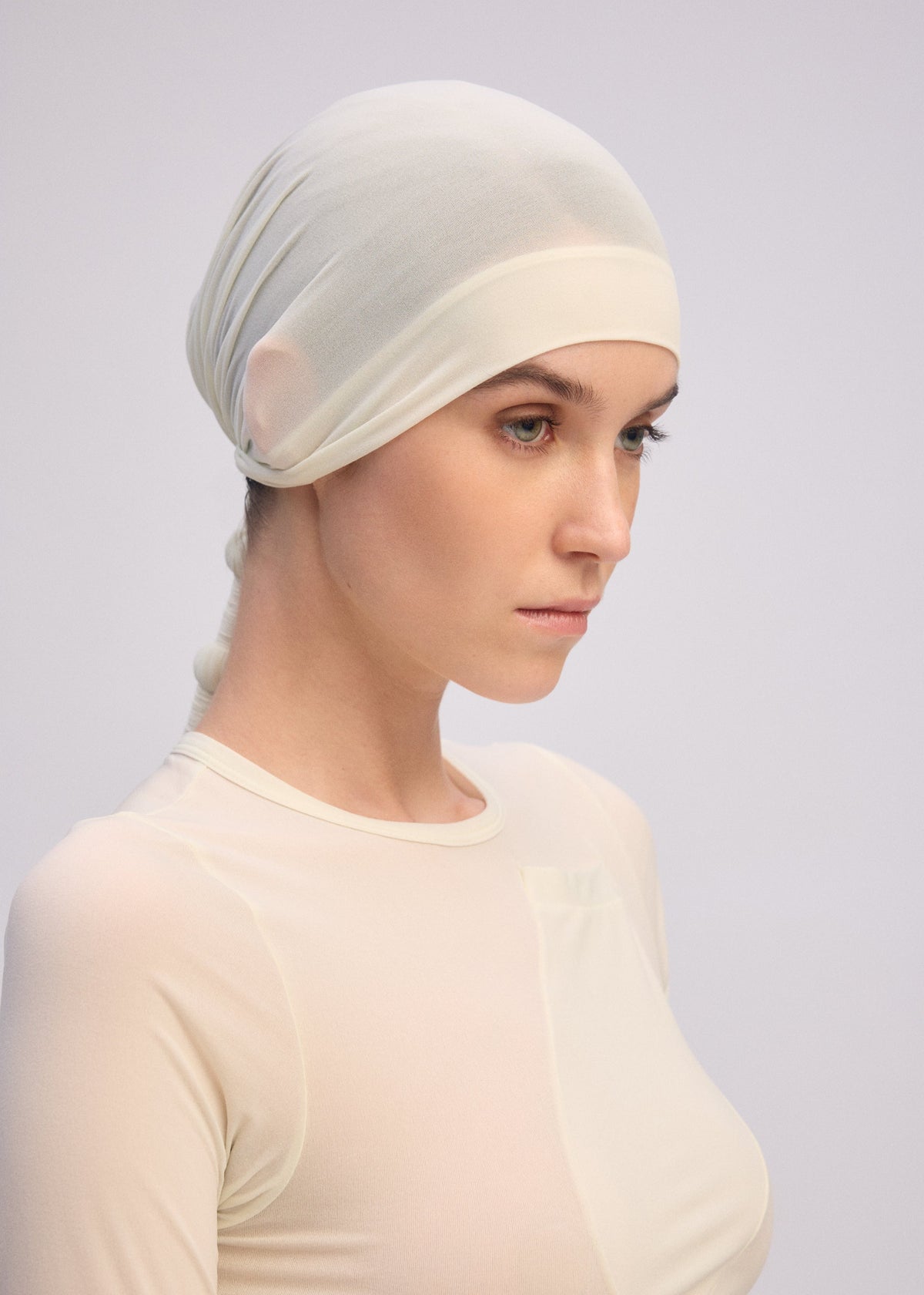A woman in the ISSEY MIYAKE WRAPPED PONYTAIL, a sheer light beige headscarf with a ponytail silhouette and matching long-sleeved top, gazes down gently against a pale neutral background, creating a soft minimalist portrait. view 6 of 7