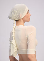 A model wears the ISSEY MIYAKE WRAPPED PONYTAIL: a sheer, cream-colored top paired with a matching, knot-styled jersey cap featuring draped fabric at the end, showcased from the back and side against a plain background.