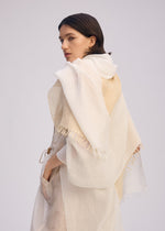 A woman with straight brown hair drapes the ISSEY MIYAKE SLITS STOLE in sheer off-white cotton with frayed edges over her shoulders, highlighting its versatile style as she gazes back against a simple light background.