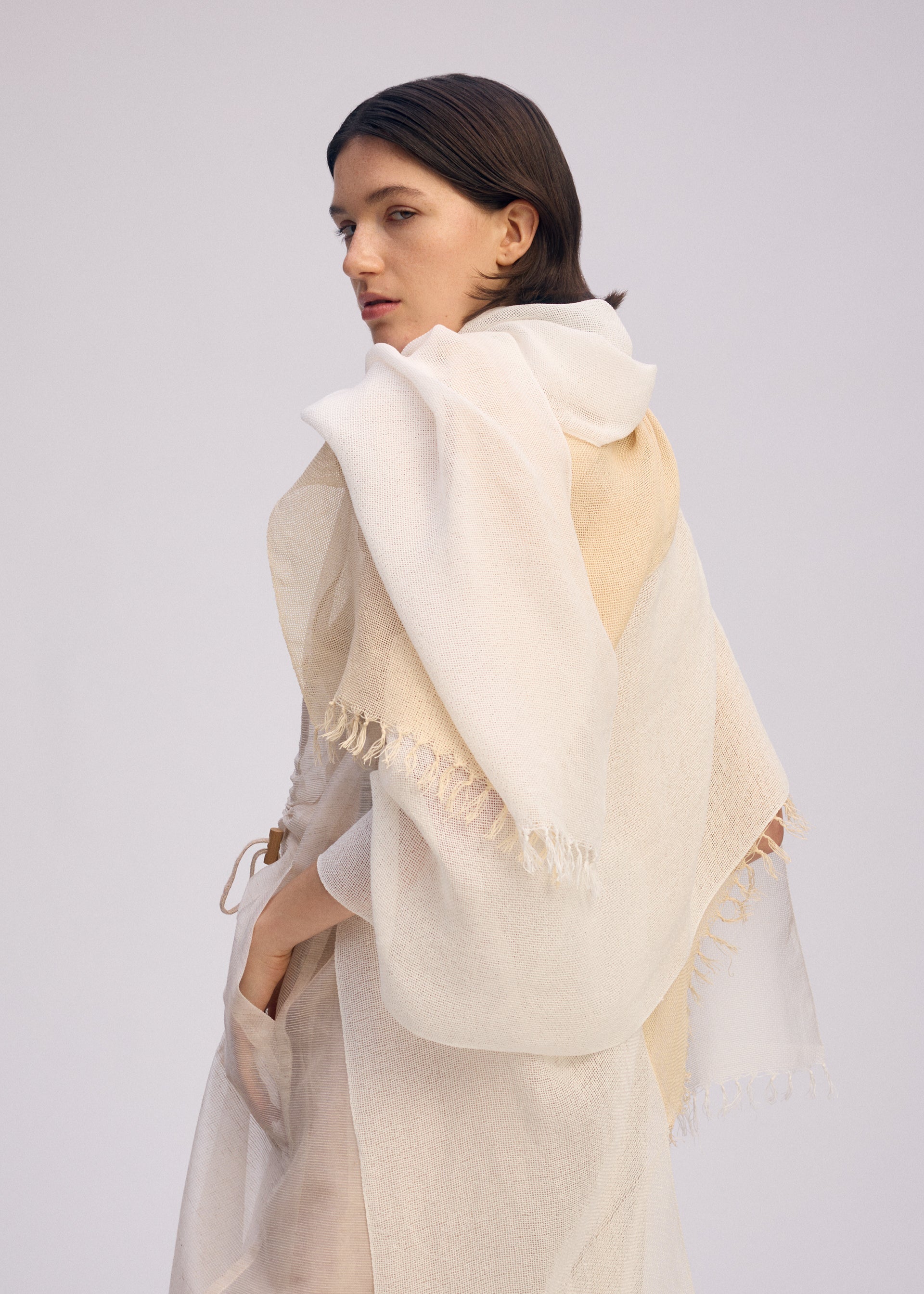 Zoomed view A woman with straight brown hair drapes the ISSEY MIYAKE SLITS STOLE in sheer off-white cotton with frayed edges over her shoulders, highlighting its versatile style as she gazes back against a simple light background.