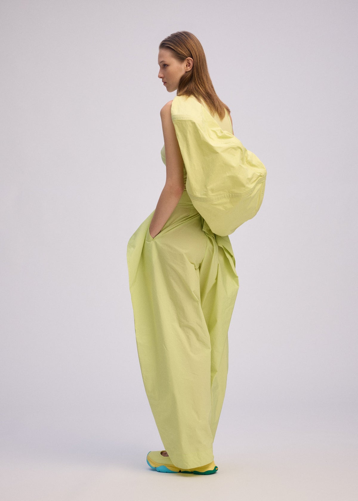 A woman stands sideways with hands in pockets, wearing ISSEY MIYAKE's PAPER BALLOON 2—a light yellow, asymmetrical, crinkled outfit—paired with turquoise shoes against a plain light background. view 5 of 6