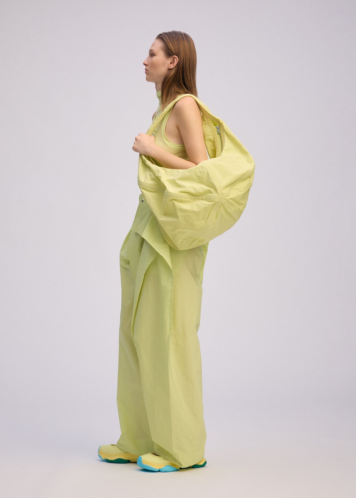 A woman stands in profile against a plain background, wearing ISSEY MIYAKE's PAPER BALLOON 2: loose, light yellow pants, a matching sleeveless top, and a large crinkled-texture shoulder bag, paired with yellow shoes with blue soles. view 6 of 6