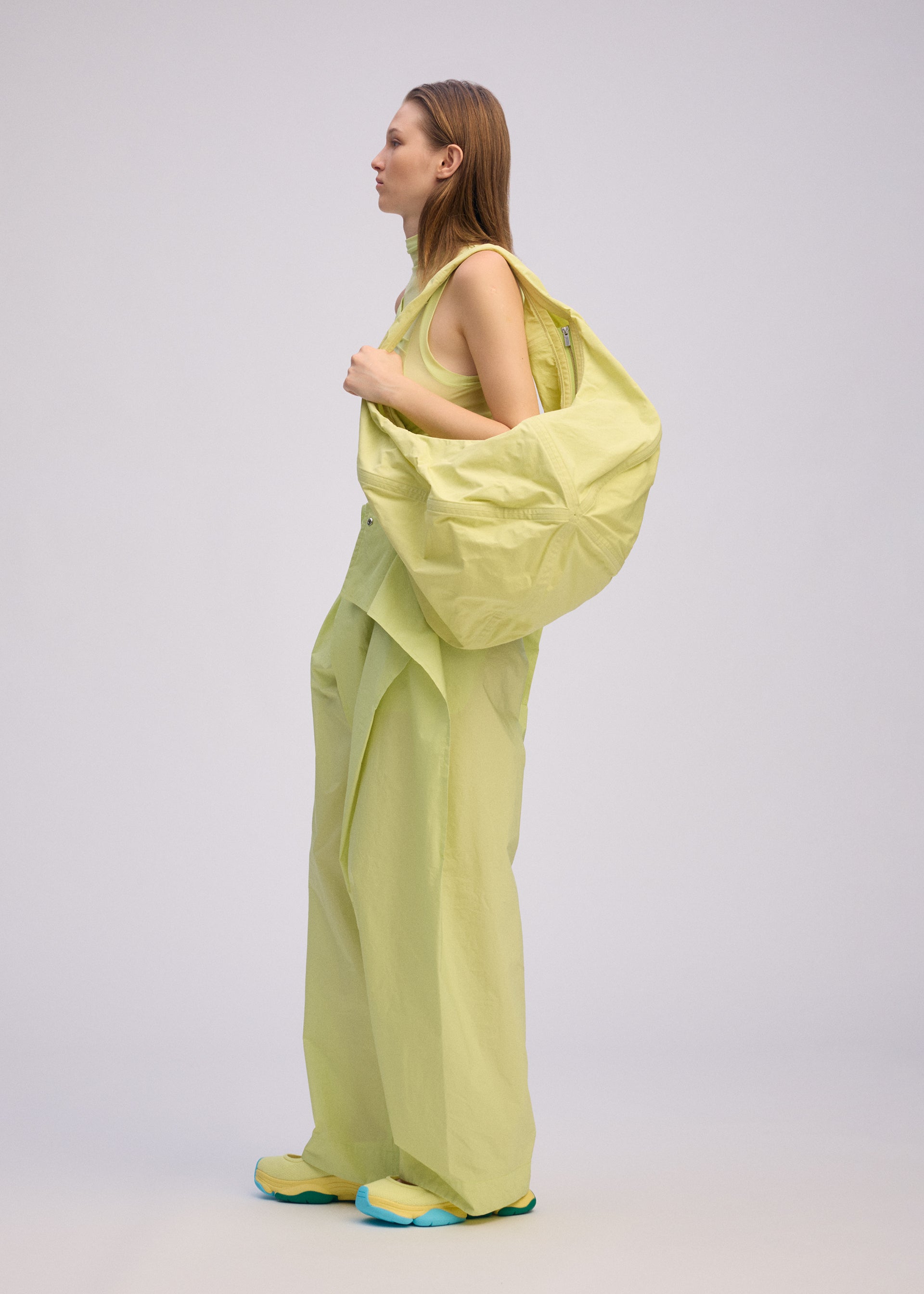 Zoomed view A woman stands in profile against a plain background, wearing ISSEY MIYAKE's PAPER BALLOON 2: loose, light yellow pants, a matching sleeveless top, and a large crinkled-texture shoulder bag, paired with yellow shoes with blue soles.