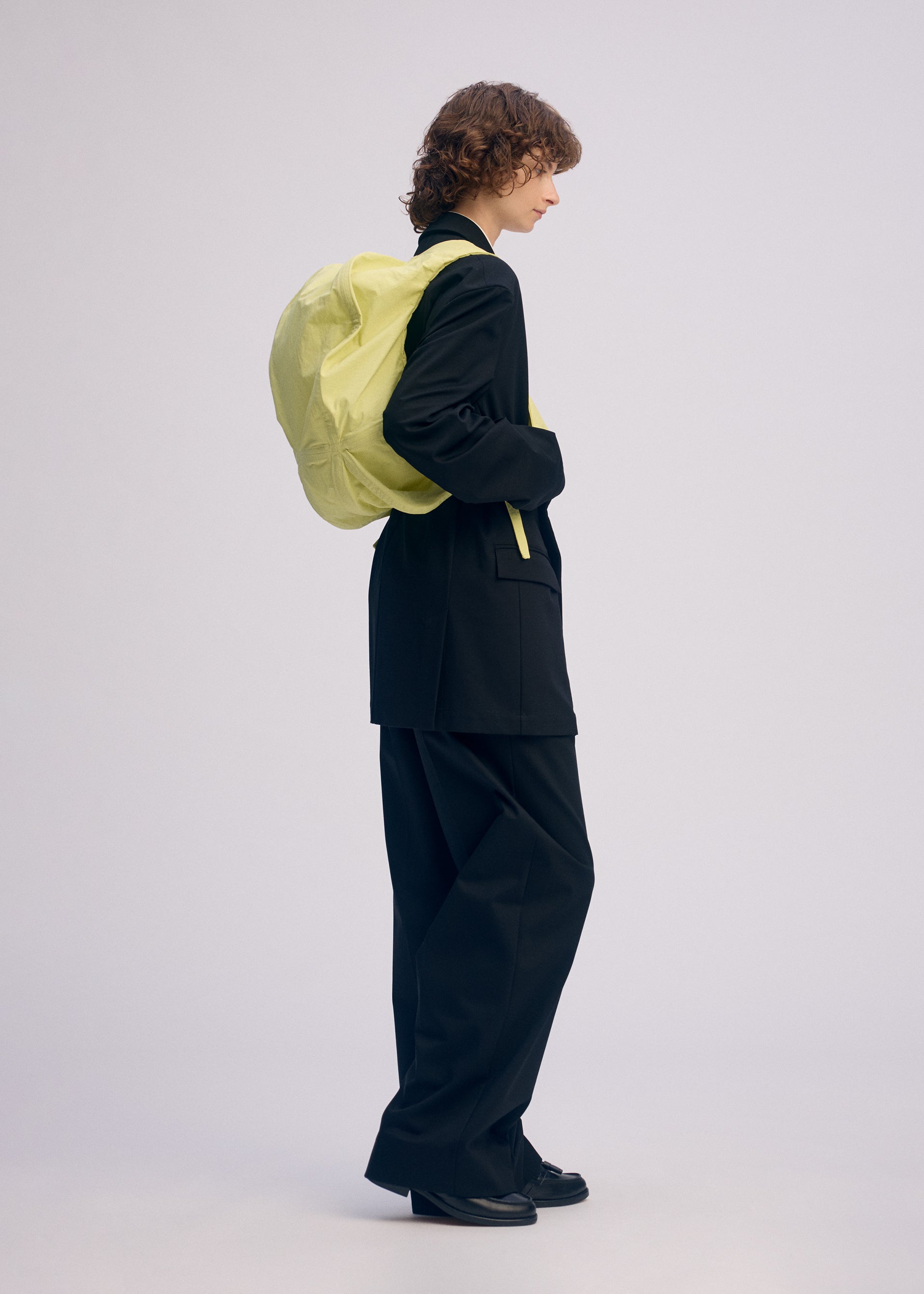 Zoomed view A person with curly hair in a loose black suit and black shoes stands in profile against a plain background, carrying ISSEY MIYAKE’s PAPER BALLOON 2 oversized light yellow backpack with a crinkled texture like Japanese Washi paper.