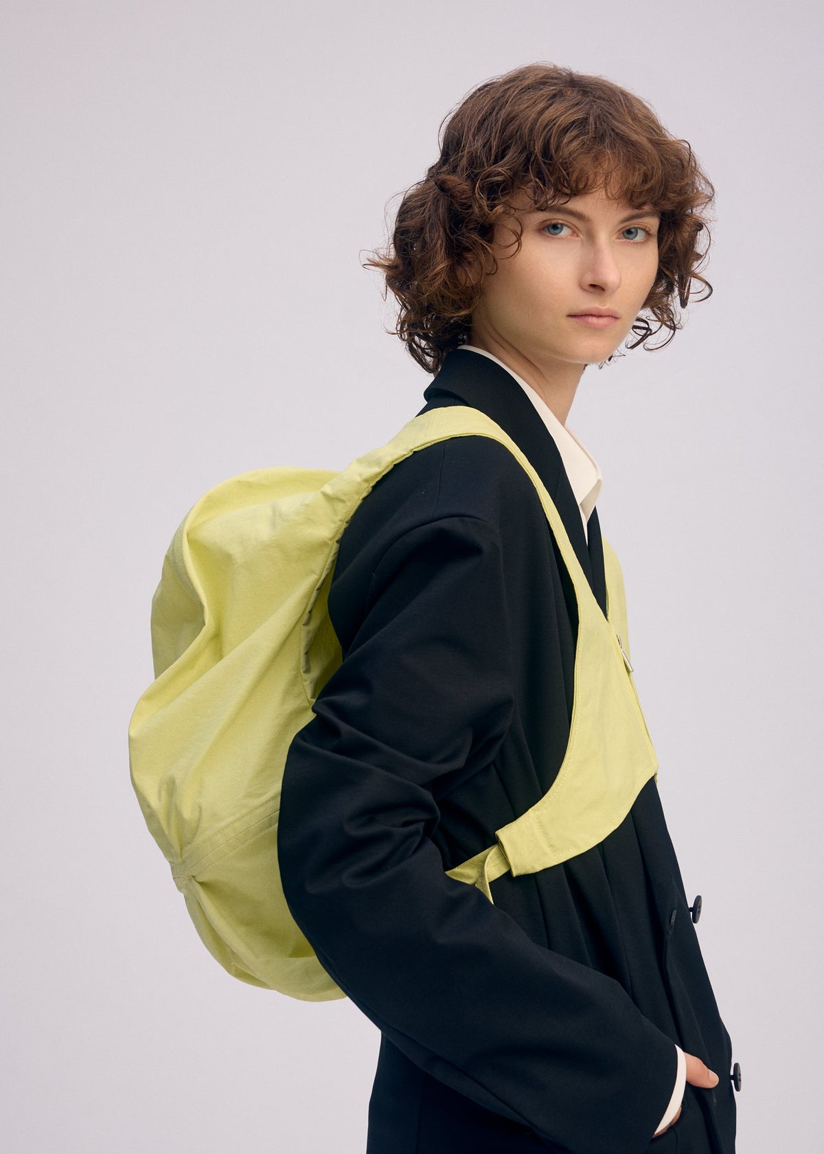 A person with short, curly brown hair wears a black blazer and white collared shirt, carrying the ISSEY MIYAKE PAPER BALLOON 2 light yellow backpack against a plain background textured like Japanese Washi paper. view 9 of 10