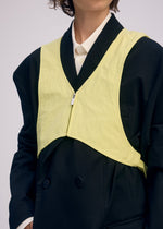 A person wears the ISSEY MIYAKE PAPER BALLOON 2 sleeveless, zippered yellow vest with a crinkled texture over a white collared shirt and loose black blazer; the cropped image shows upper torso and part of the face.