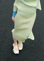 A person stands on asphalt, showing only their lower half, dressed in a light green ribbed skirt and ISSEY MIYAKE KARST FINCH sneakers with colorful blue accents.