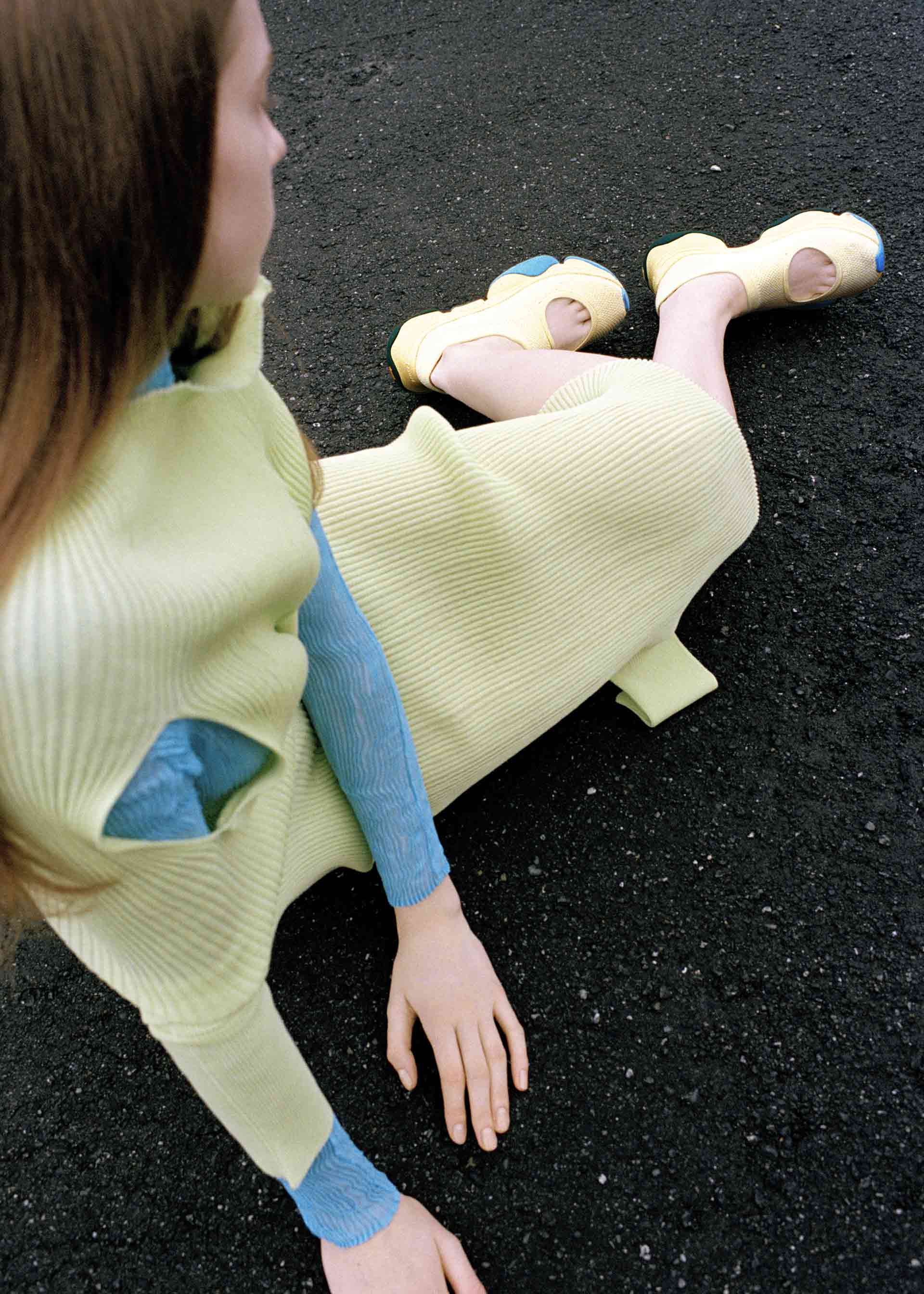 Zoomed view A person with long brown hair sits on dark asphalt, wearing a pale yellow ribbed dress over a blue top and matching ISSEY MIYAKE KARST FINCH sneakers. The relaxed pose is captured from above.
