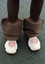 A person wearing wide, ribbed brown pants with long, flared legs that cover most of their ISSEY MIYAKE KARST FINCH sneakers, standing on a textured asphalt surface.