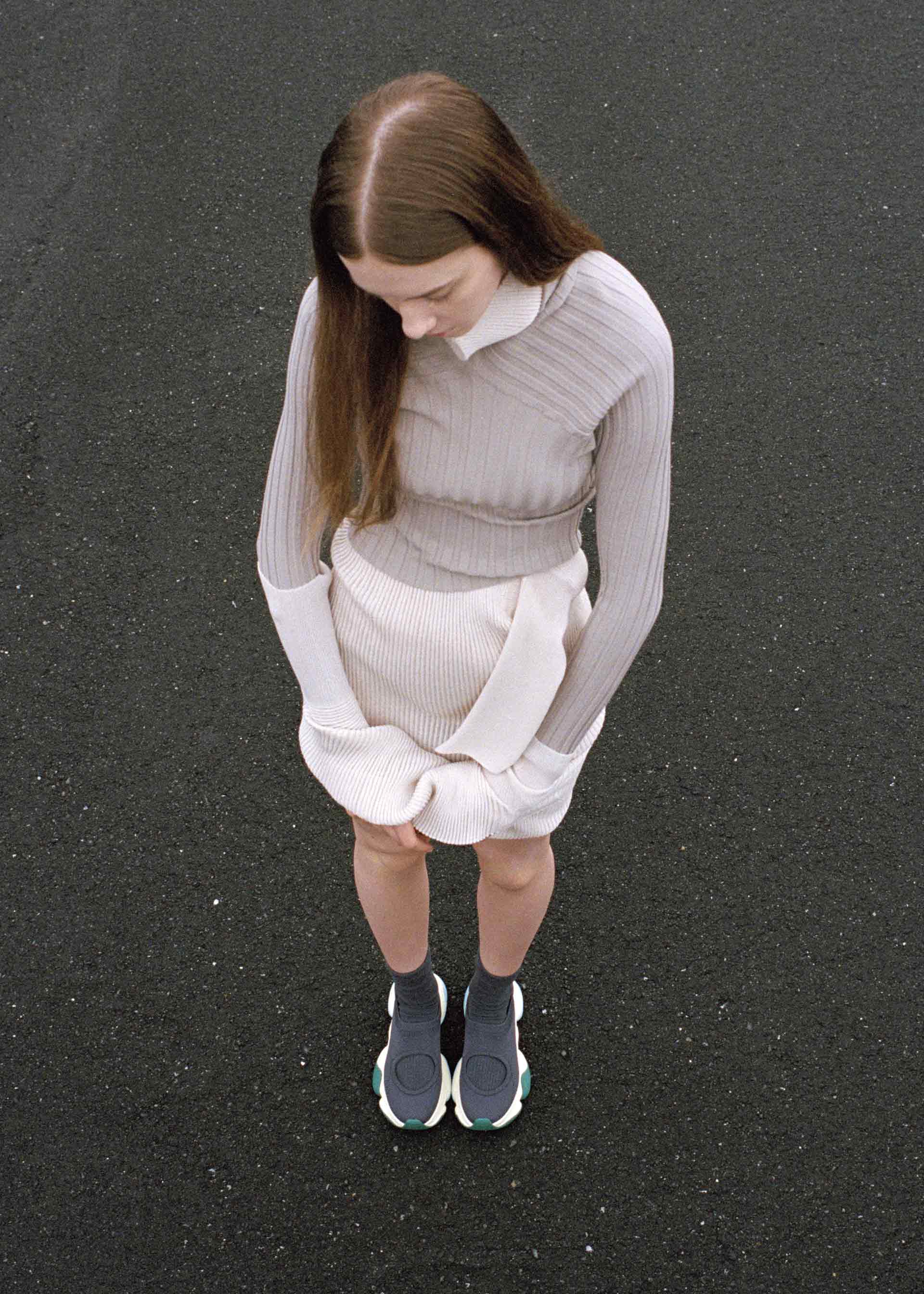 Zoomed view A woman with long brown hair stands on a dark pavement, looking down. She wears a light, ribbed long-sleeve dress and unique ISSEY MIYAKE KARST FINCH sneakers, adding colorful, lightweight style to her futuristic look.